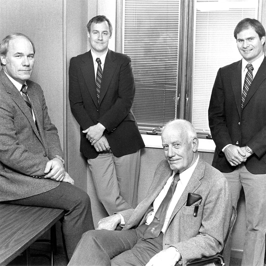 black and white photo of four adult men wearing suits posing inside an office